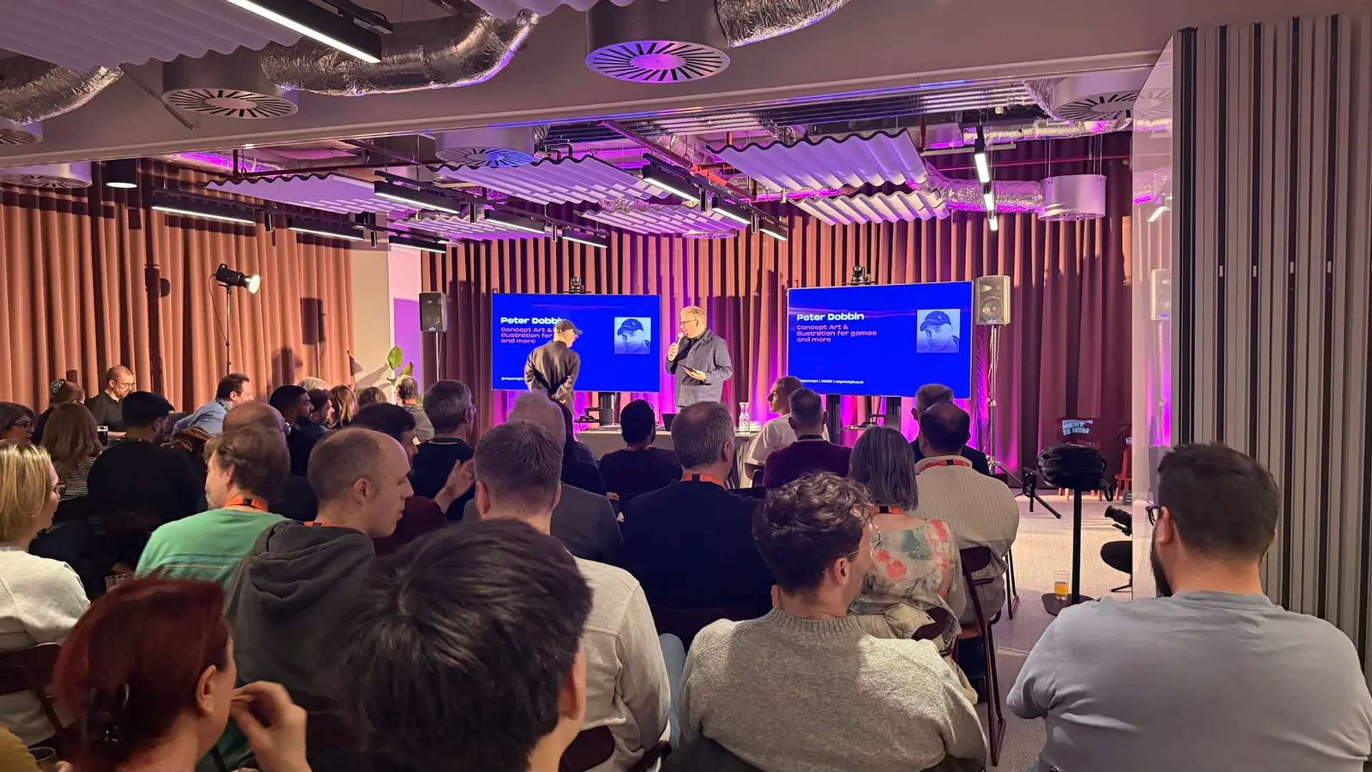 The stage at MKGN, with the host and a speaker in front of two screens showing a Peter Dobbin slide, "Concept Art & illustration for games and more", and a seated audience in the foreground